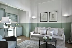 Un salon moderne avec un canapé, une table basse et une lampe. On y trouve également un coin cuisine équipé d’un réfrigérateur et d’une étagère où sont rangés des verres.
