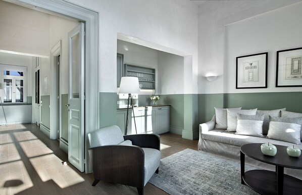 Un salon moderne avec un canapé, un fauteuil et une table basse. On aperçoit un coin cuisine avec des placards et une table à manger à travers une porte ouverte.