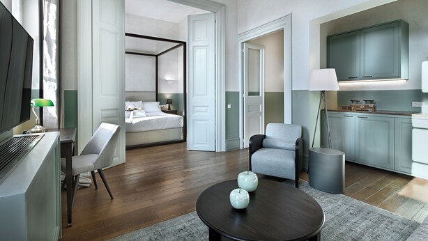 A modern hotel room with a bed, a seating area with a table, and a kitchenette.