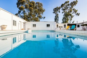 A swimming pool with clear blue water, surrounded by a white building with windows and doors, and trees in the background.