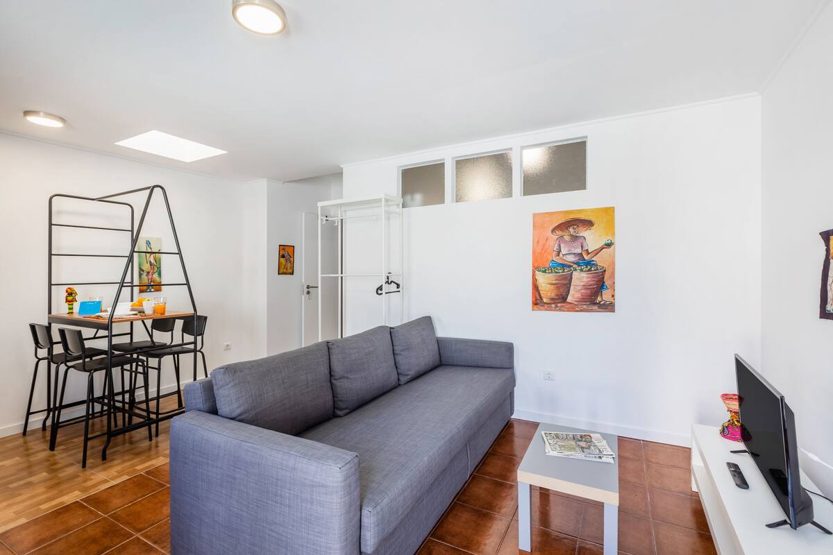 A modern living room with a grey sofa, a dining table with chairs, a TV, and artwork on the walls.