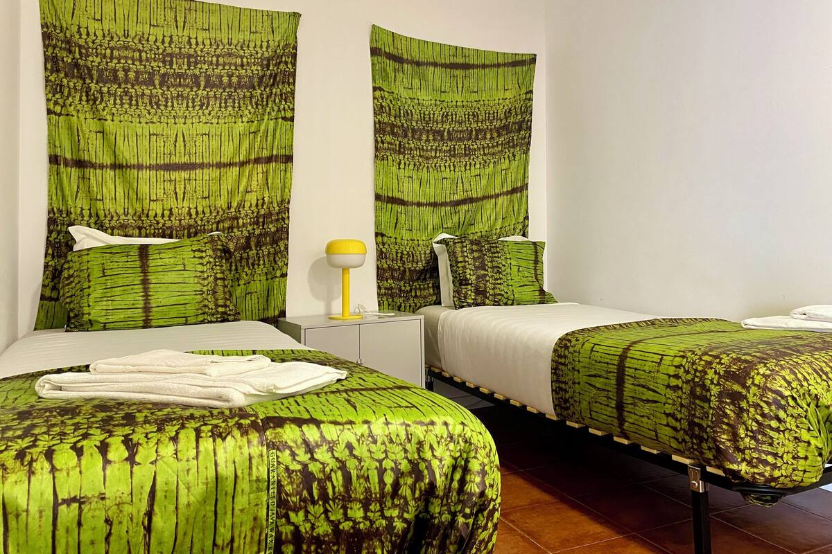 A hotel room with two beds, each with green patterned bedding, white pillows, and a yellow bedside lamp.