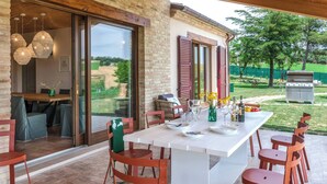 Ein Essbereich im Freien mit einem weißen Tisch und roten Stühlen, einer Ziegelwand und Blick auf Garten und Grillplatz.
