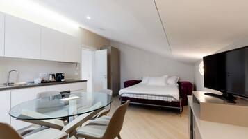A modern hotel room with a glass dining table, a bed with a red headboard, a TV, and a kitchenette.