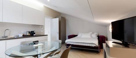 A modern hotel room with a glass dining table, a bed with a red headboard, a TV, and a kitchenette.