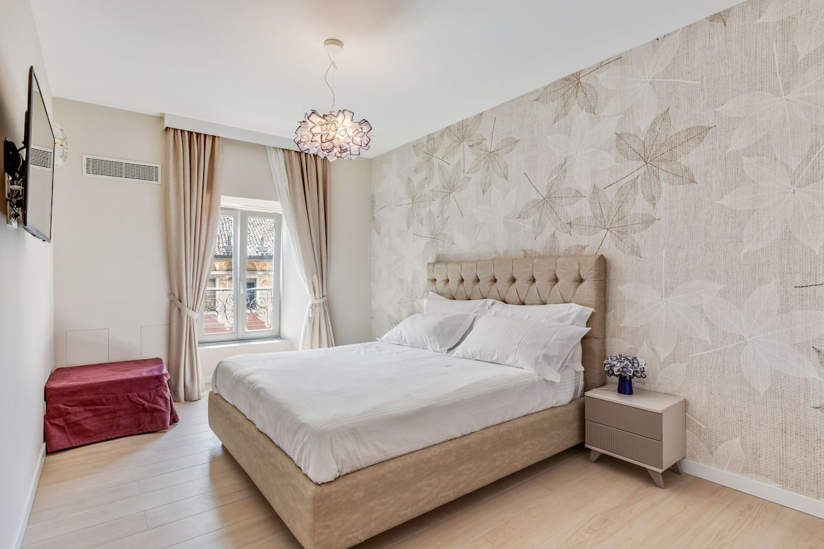 Ein Schlafzimmer mit einem großen Bett, einem Nachttisch, einem Spiegel und einem Fenster mit Vorhängen.