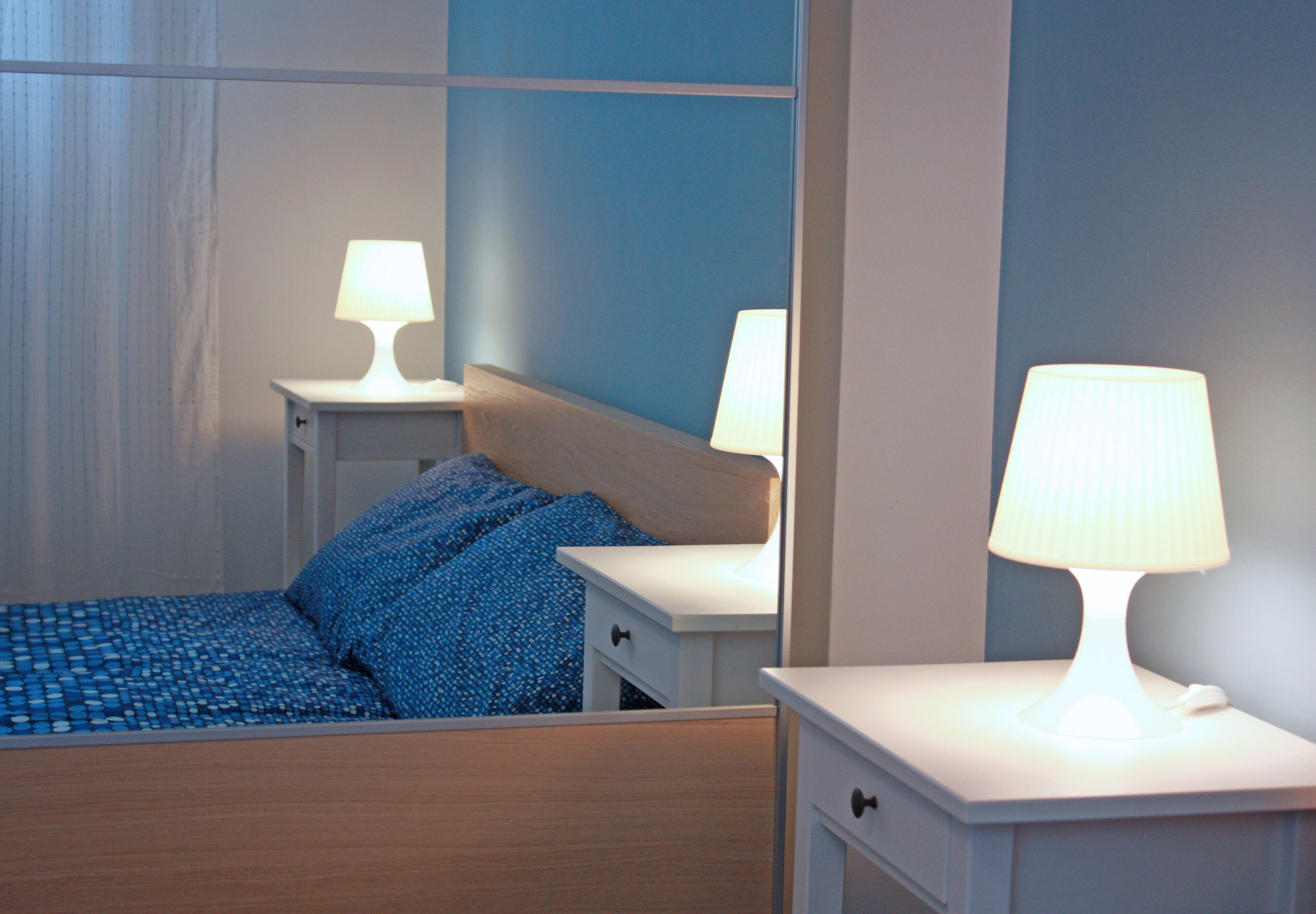 Cama bem arrumada com roupa de cama azul, duas mesas de cabeceira com abajures e uma mesinha de cabeceira.