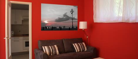 Une pièce avec un mur rouge, un canapé marron orné de coussins à rayures, une petite table blanche et un tableau représentant un coucher de soleil.