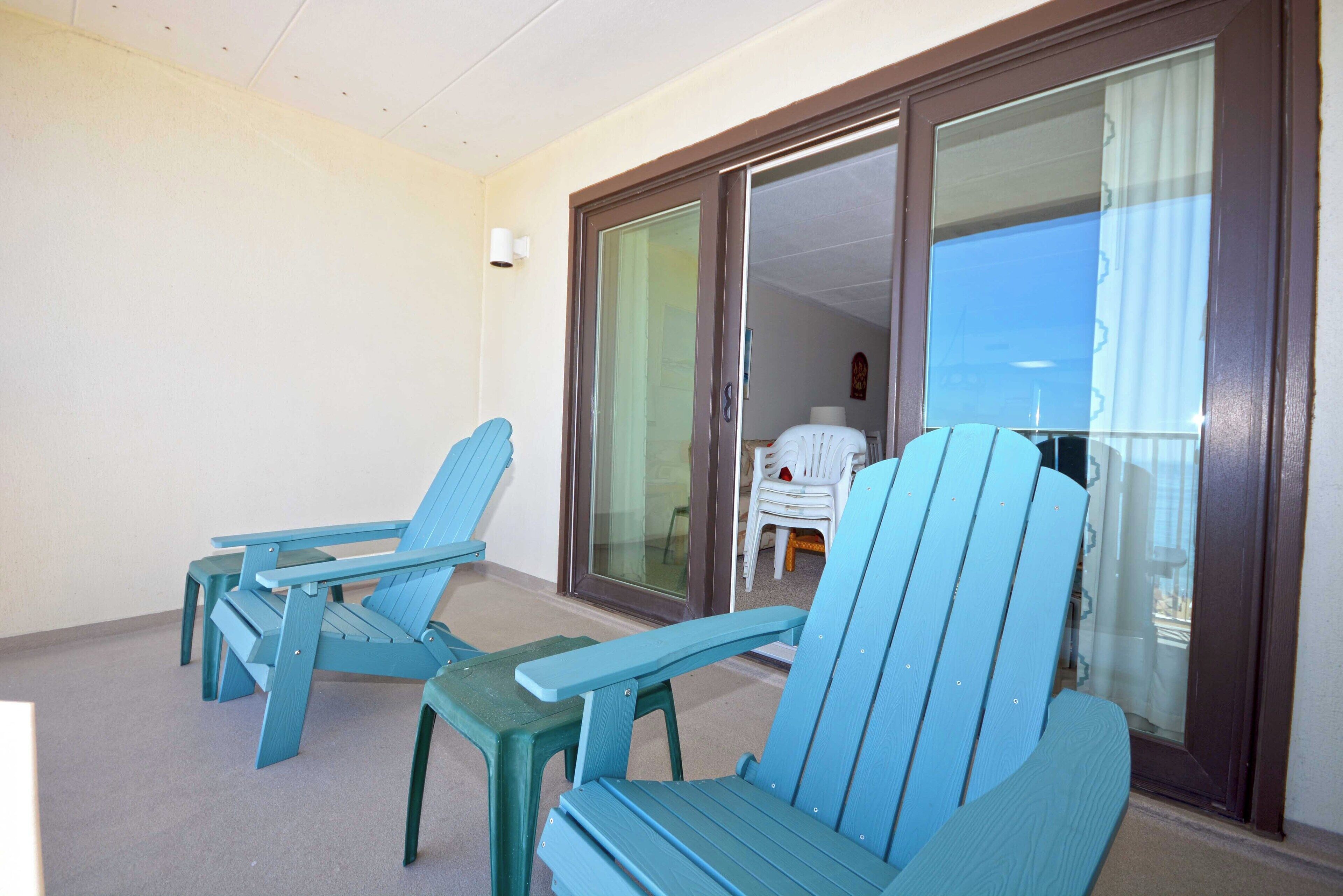 A balcony with two blue Adirondack chairs and a small table, a sliding glass door, and a view of the sky.