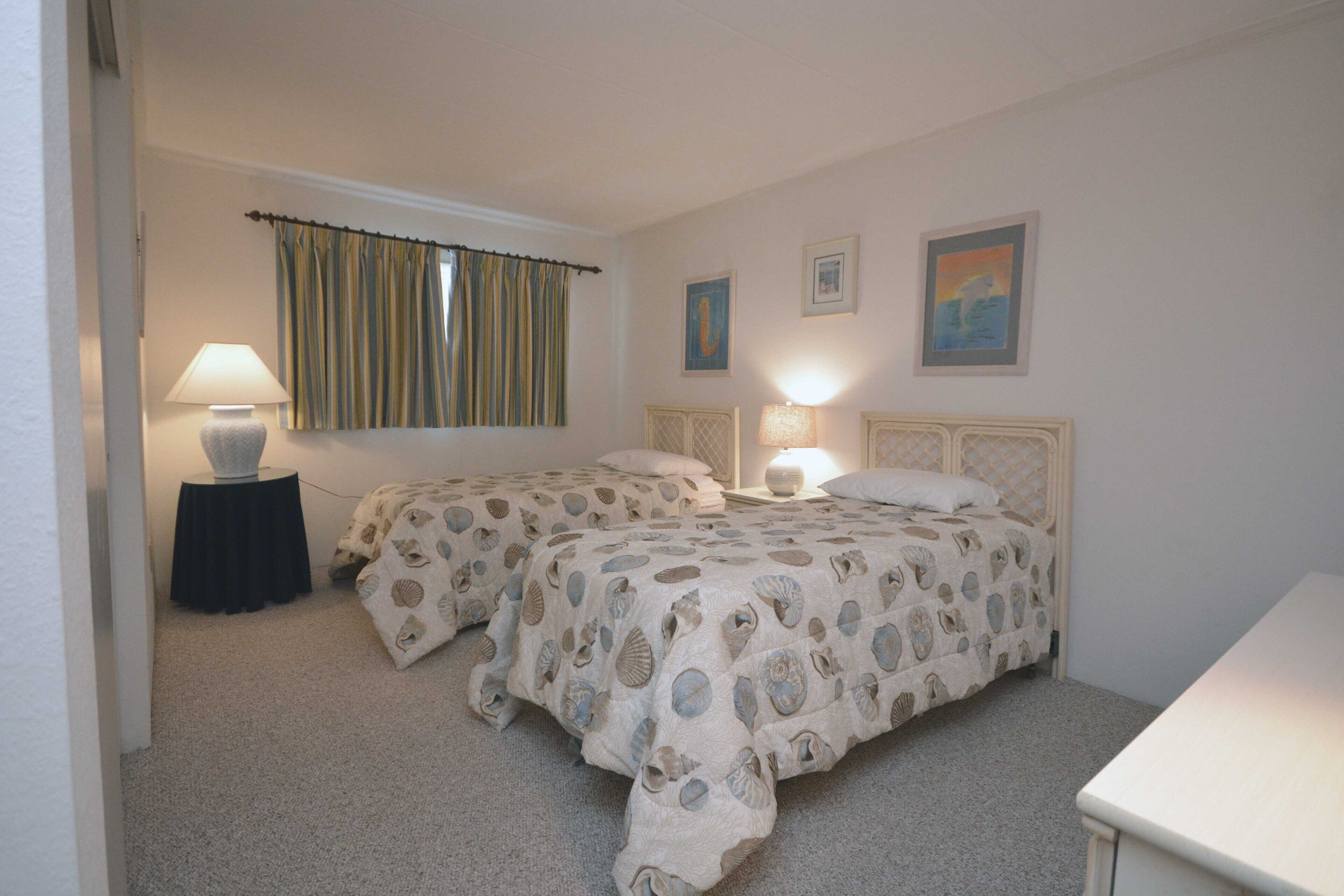 A bedroom with two beds, a window with curtains, bedside lamps, and framed pictures on the wall.