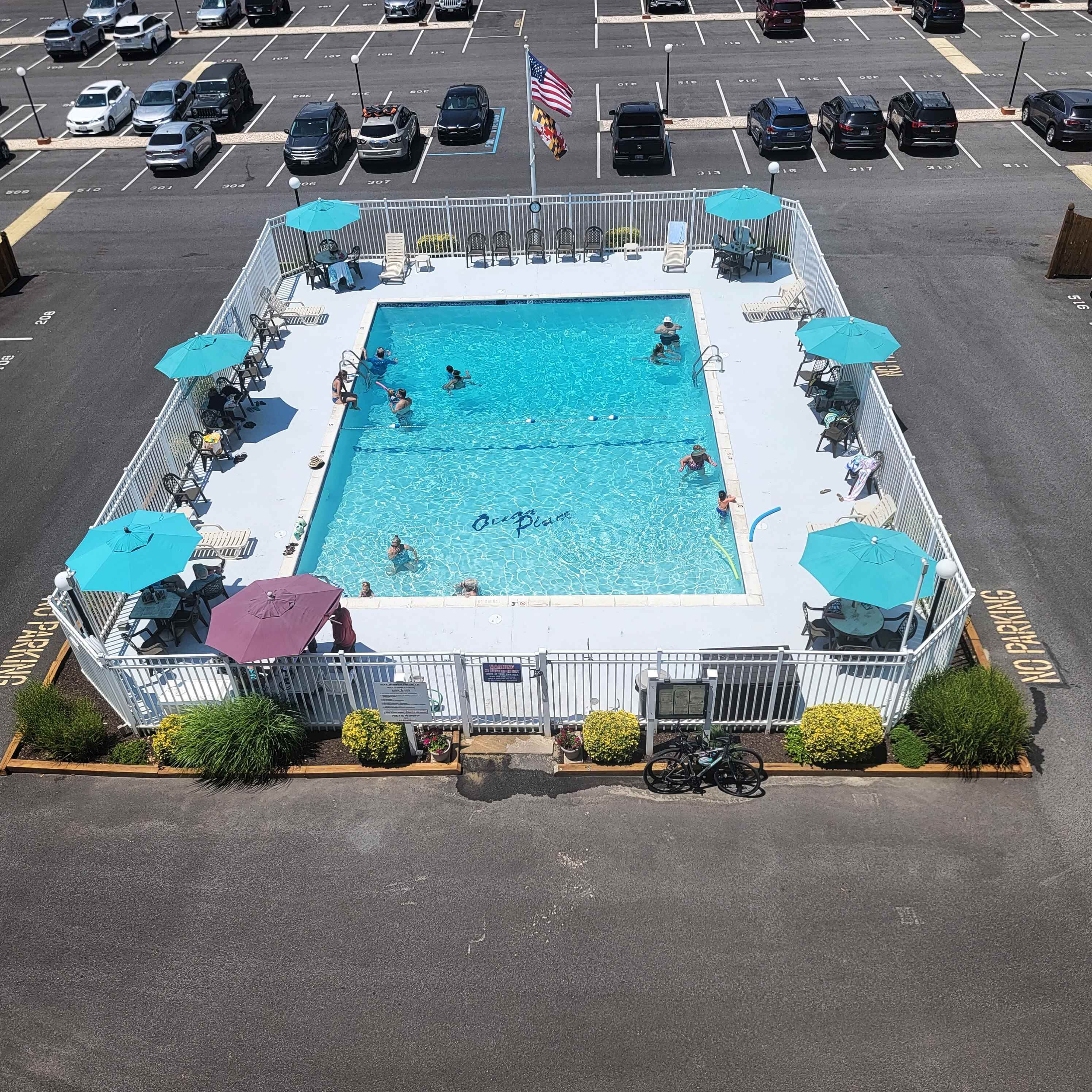 A square swimming pool with a white fence, surrounded by lounge chairs and umbrellas.