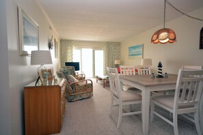Een eethoek met een houten tafel en witte stoelen, een rieten bank en een televisie op de achtergrond.