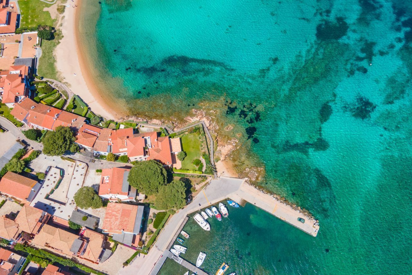 An aerial view of a coastal town with a beach, a marina with boats, and residential buildings with red roofs.
