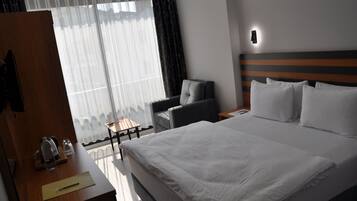Habitación de hotel con una cama grande, un escritorio con una silla, una mesita de noche y una ventana con cortinas traslúcidas.