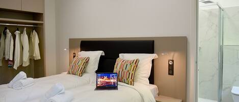 A modern hotel room with a bed, a laptop, a shower, and a wardrobe with hanging clothes.