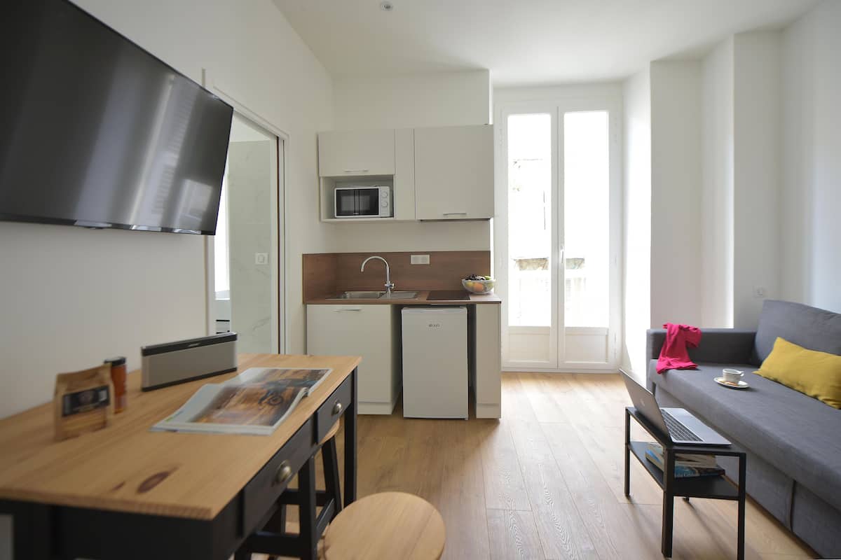 A compact kitchen with a microwave, sink, and a compact refrigerator. There's a dining table with a laptop and a book, and a sofa with a yellow pillow.