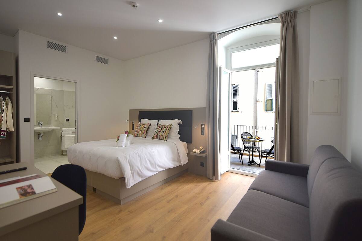 A hotel room with a bed, a desk, a chair, a sofa, a bathroom, and a balcony with a table and chairs.