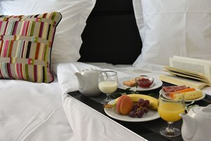Una bandeja de desayuno ordenada sobre una cama, con un almohadón, un libro y una bandeja de comida que incluye frutas, queso y huevos.