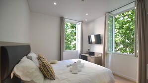 Ein Hotelzimmer mit einem Bett, einem Schreibtisch mit Computer, einem Fernseher und großen Fenstern mit Vorhängen.