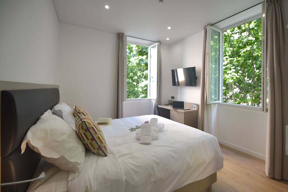 A hotel room with a bed, a desk with a computer, a TV, and large windows with curtains.