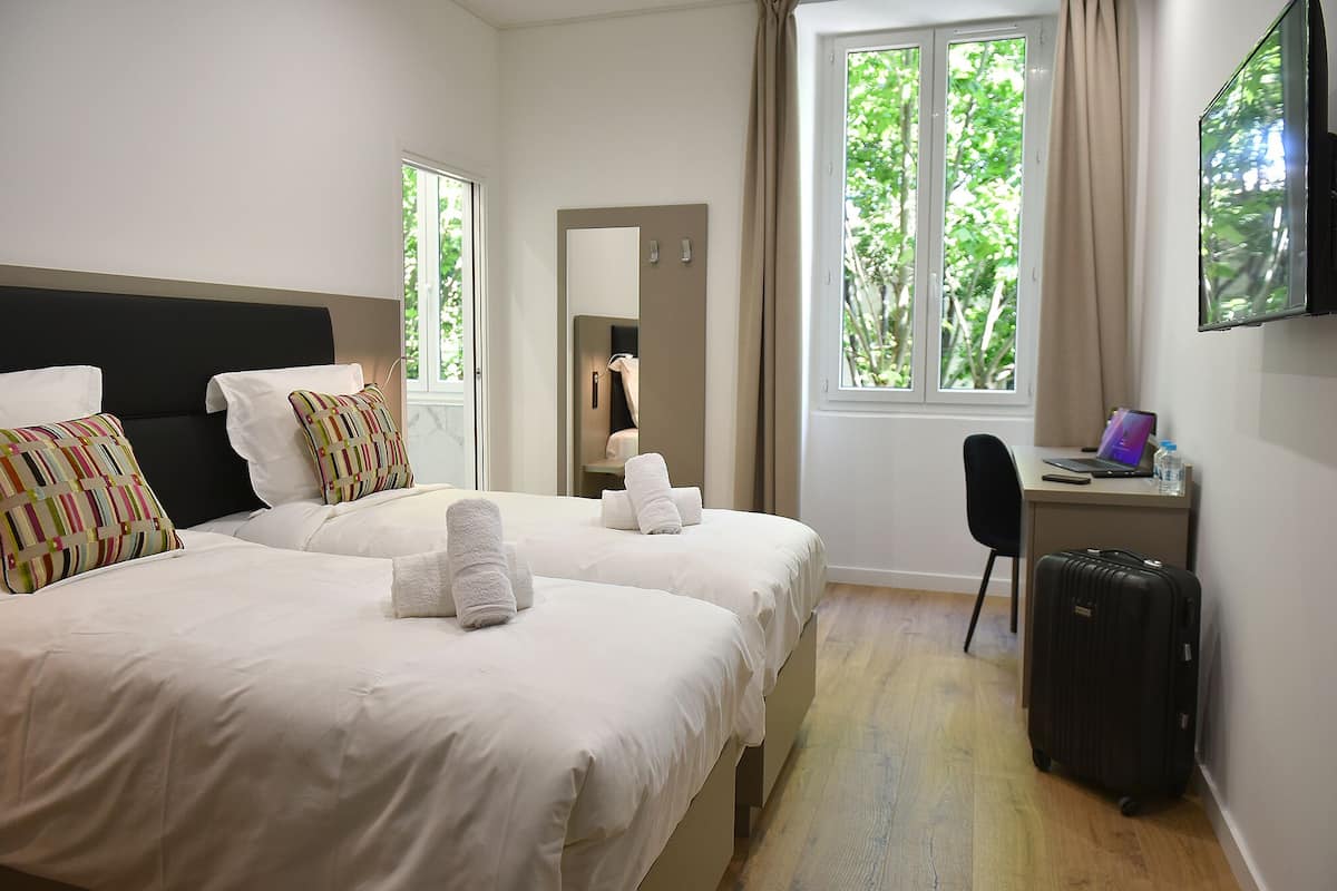 A hotel room with two beds, a desk, a chair, a TV, and a window with a view of trees.