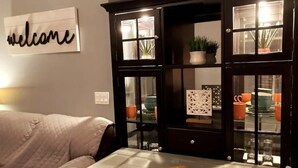Een woonkamer met een bank, een tafel met onderzetter, een kast met glazen deuren en een bord met de tekst 'welkom' aan de muur.