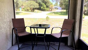 Een hoekzaak met twee stoelen en een kleine tafel, een raam met uitzicht op een woonstraat en groen.
