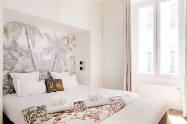 A hotel room with a large bed, a headboard with a palm tree design, and a window with a view of buildings. - A Breathtaking 2BR/2BA in Le Marais (Paris)