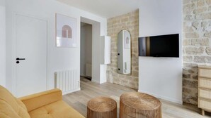 A modern living room with a sofa, two wicker ottomans, a stone wall, a mounted TV, and a white door. - A Chic 1-BR in Sentier - Montorgueil (Paris)