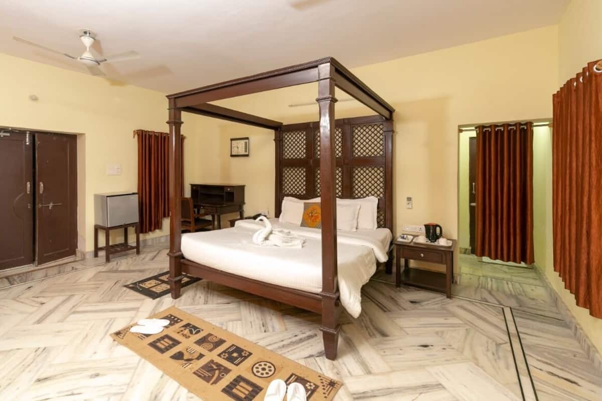 Uma cama com dossel, colchão branco e travesseiro decorado com cisne, mesa de cabeceira de madeira com luminária, televisão em um suporte e piso com padrão herringbone.