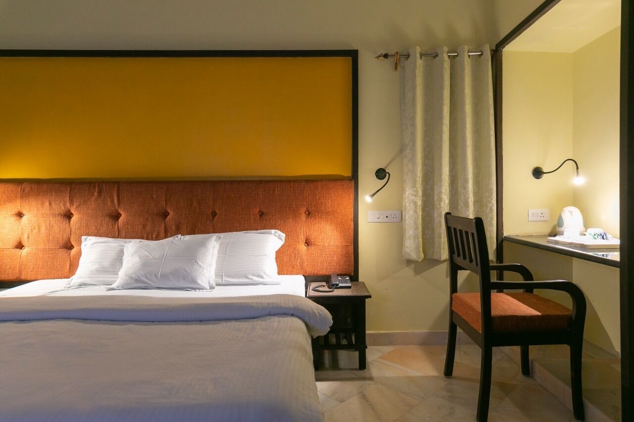 A hotel room with a large bed, a wooden headboard, a bedside table with a lamp, a chair, and a desk with a mirror and a telephone.