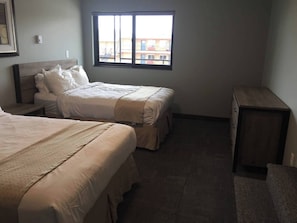 A hotel room with two beds, a nightstand, a dresser, and a window with a view of buildings. - Suite 213 - 1BR with 2 beds, kitchen, 2nd floor (Rapid City)
