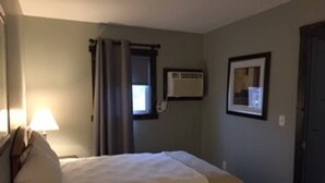 A hotel room with a bed, a bedside lamp, a window with curtains, a framed picture on the wall, and a door.