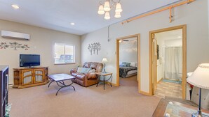 A spacious living room with a leather sofa, a TV, and a wooden cabinet. There is a window with blinds, a lamp, and a door leading to another room. - Large 1BR Condo Near Lake Pepin - Starshine (Pepin)