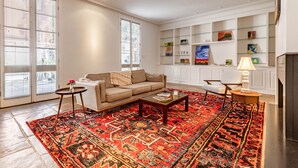 Una sala de estar con un sofá color beige, una mesita de centro, un tapete rojo estampado y una estantería con varios objetos.