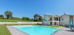 Une piscine entourée d’une zone dallée, un bâtiment à deux étages doté de balcons et un paysage verdoyant avec des arbres.