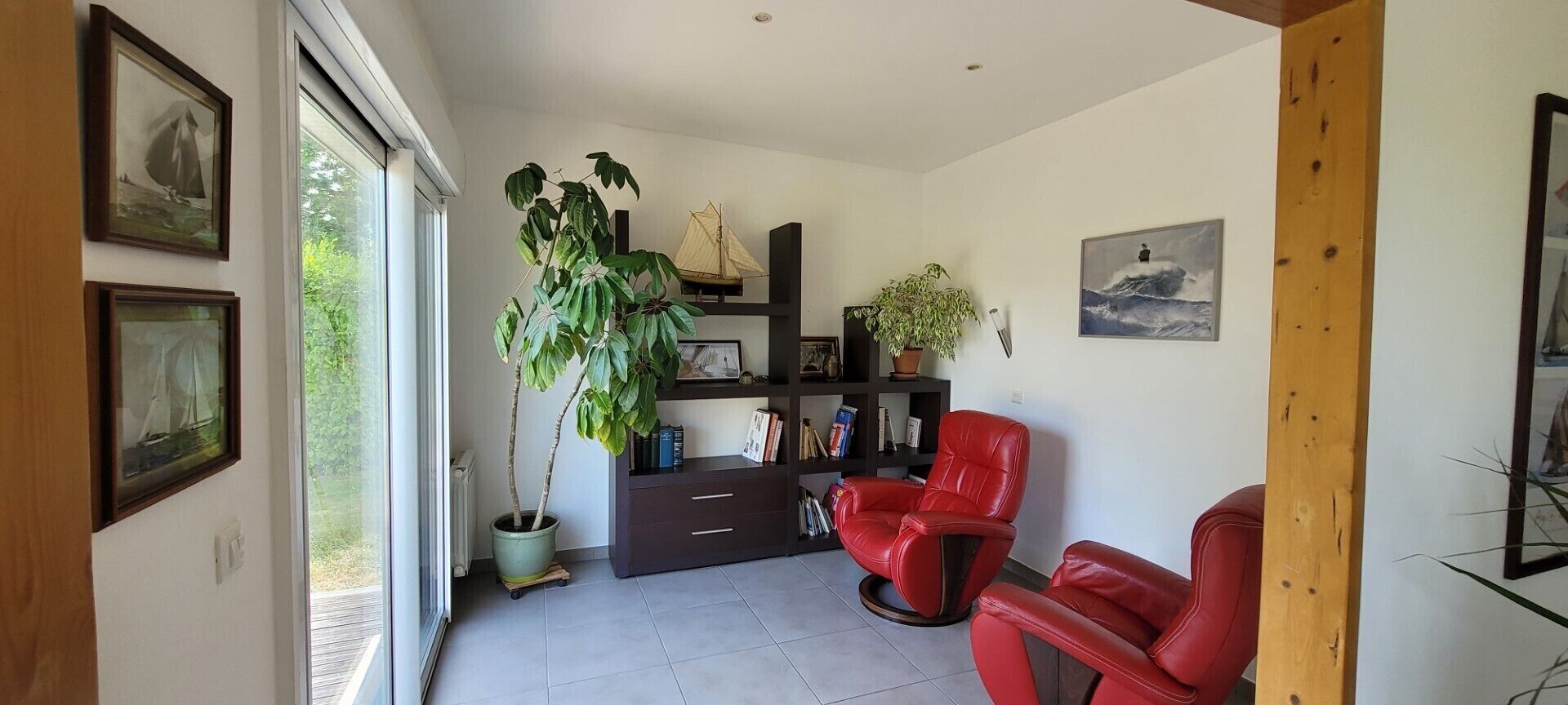 Un moderno salón con un ventanal grande, una planta en maceta, un sillón de cuero rojo, una estantería y un cuadro en la pared.