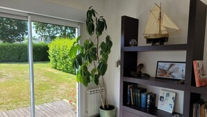 Une pièce dotée d’une porte-fenêtre coulissante donnant sur un jardin, d’une bibliothèque remplie de livres et d’une maquette de bateau, ainsi que d’une plante en pot.