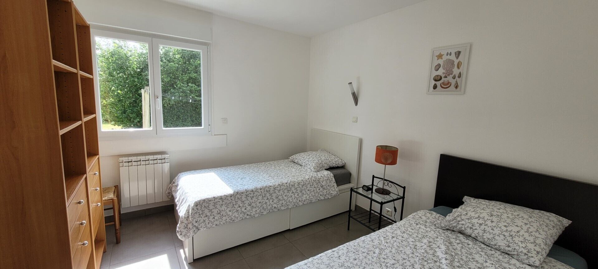 A bedroom with two beds, a radiator, a window, and a wall-mounted lamp.