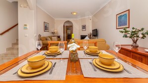 A dining area with a wooden table set for four, a vase with flowers, and a sofa in the background.