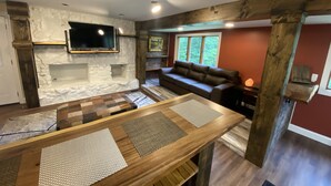 Een gezellige woonkamer met een stenen openhaard, een houten tafel met plaatsmatten, een leren bank en een groot raam met uitzicht op bomen.