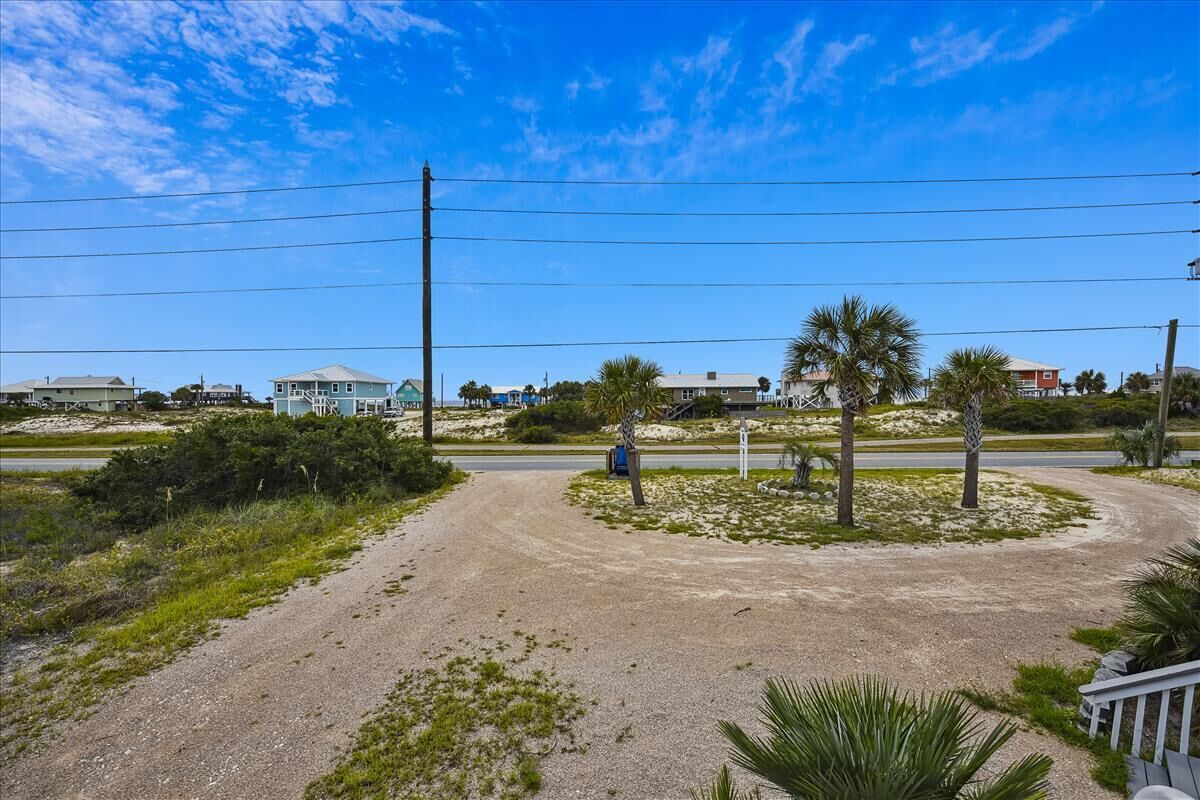 A coastal road with a dirt path, palm trees, and houses in the background.