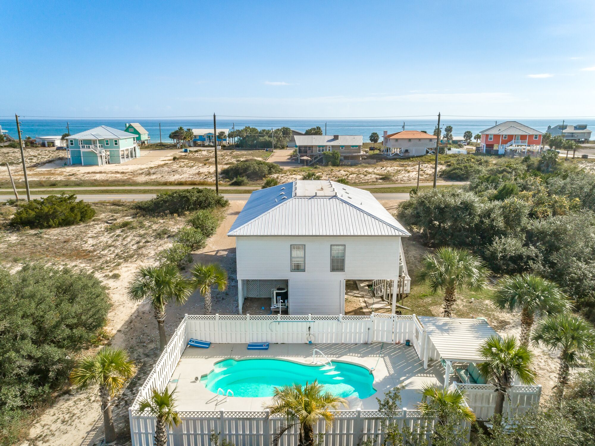 A house with a swimming pool surrounded by a fence, located near a beach with other houses in the background.