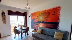 A living room with a sofa, dining table, chairs, and a mural of a tree and bird. - Stunning Ocean View! Communal Pool and Quick Walk to the Beach (Portimão)