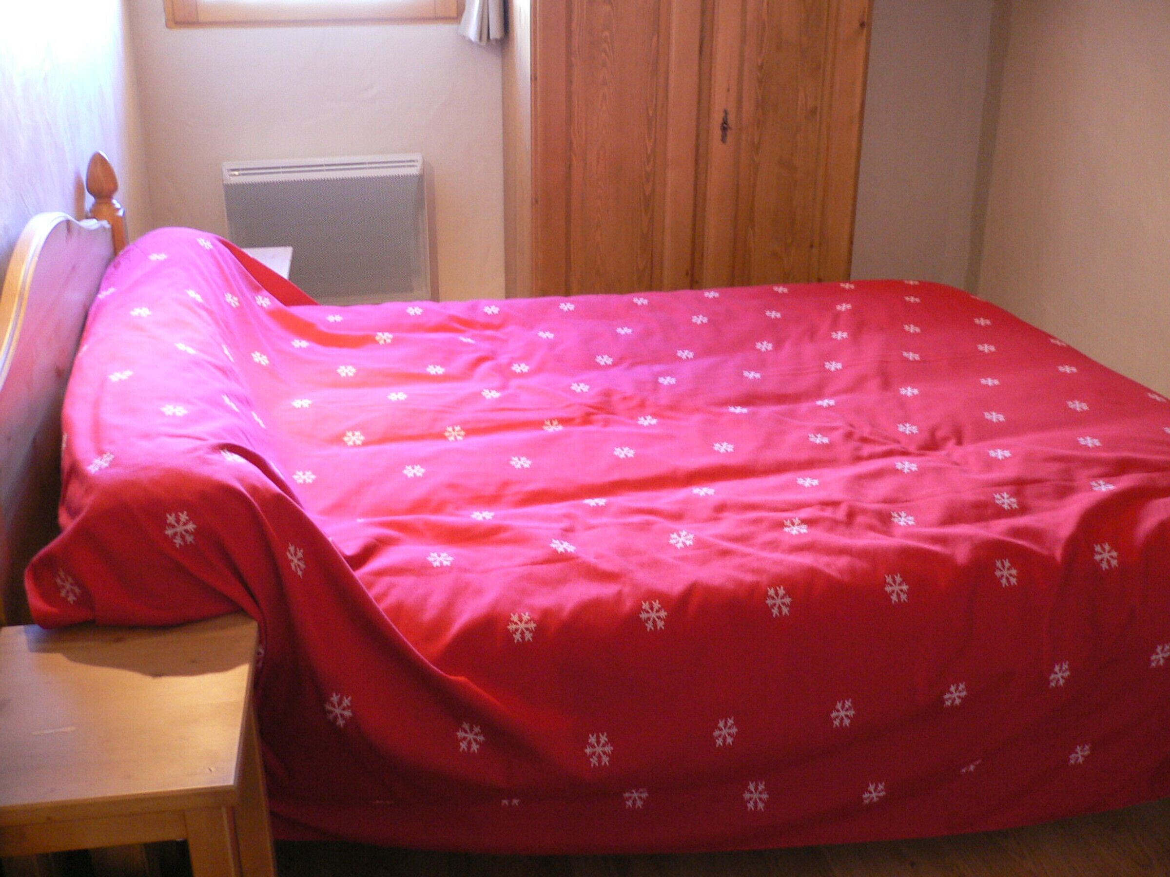 Una camera da letto con un letto ricoperto da una coperta rossa con motivi a fiocchi di neve, una testiera in legno, un comodino in legno e un termosifone sulla parete.
