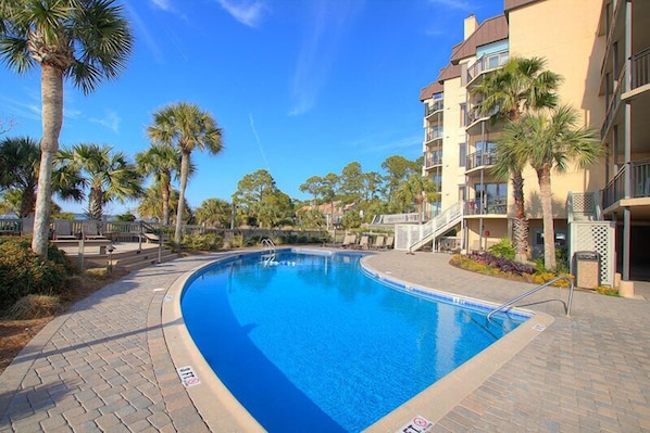 A swimming pool with a clear blue water, surrounded by palm trees and a multi-story building with balconies.