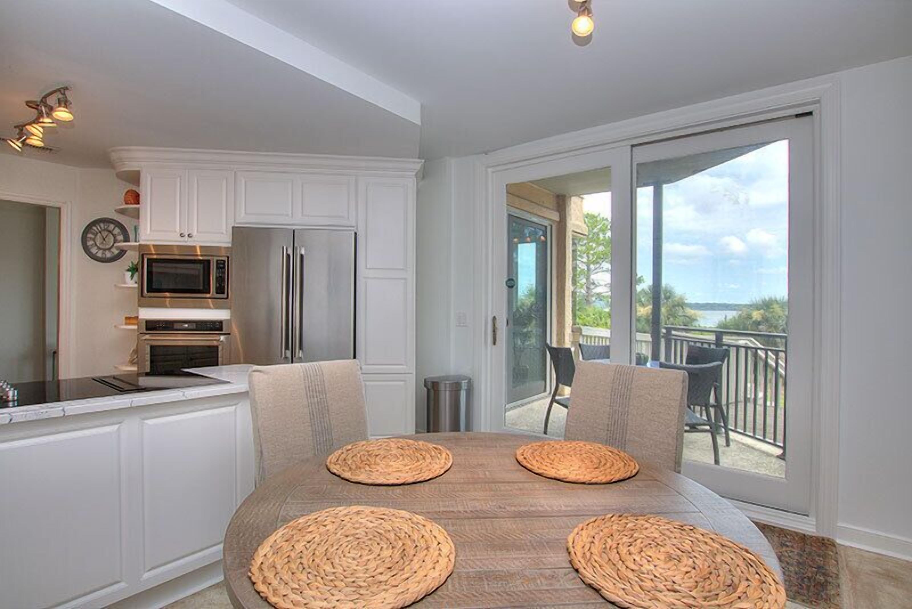 A modern kitchen with white cabinetry, a stainless steel refrigerator, and a microwave. There is a dining table with woven placemats and a view of a balcony with outdoor furniture.