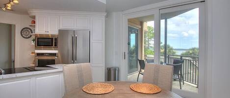 A modern kitchen with white cabinetry, a stainless steel refrigerator, and a microwave. There is a dining table with woven placemats and a view of a balcony with outdoor furniture.