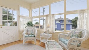 A cozy room with wicker furniture, a patterned rug, and large windows offering a view of the neighborhood.