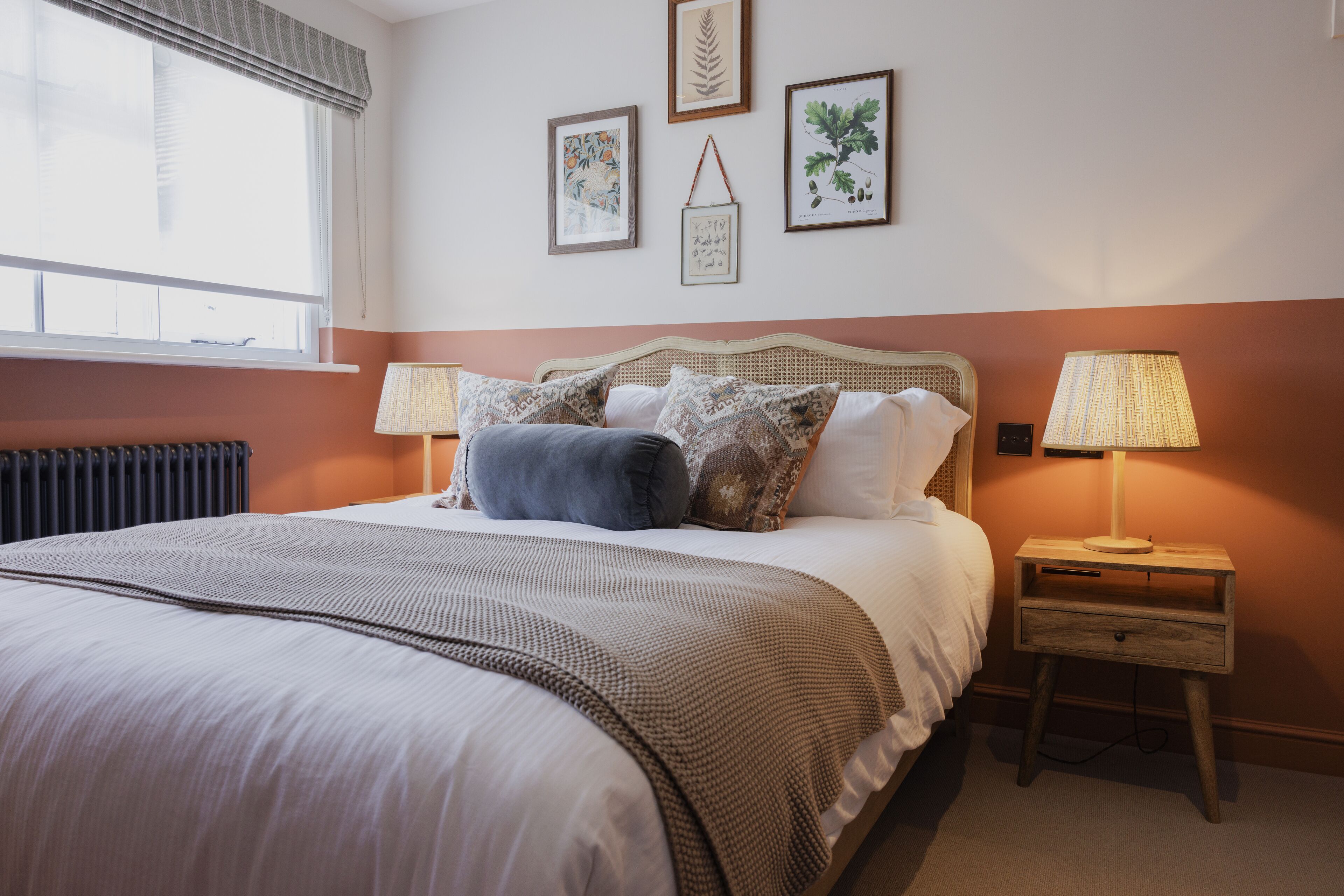 A neatly made bed with a patterned bedspread and pillows, a bedside table with a lamp, and framed botanical prints on the wall.